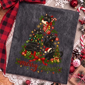 K&ouml;nnte beinhalten: Ein dunkelgraues T-Shirt mit einer Grafik aus schwarzen Katzen in Form eines Weihnachtsbaums, verziert mit Ornamenten, Lichtern und einem Weihnachtsstrumpf. Der Text "merry catmas" steht in roter und gr&uuml;ner Schrift unten.