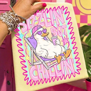 Op de afbeelding: Een lichtgele handdoek met een cartoon eend met een hartvormige zonnebril en een roze pet, ontspannen in een strandstoel. De handdoek heeft de tekst "Sizzlin' But Chillin'" in roze en paars. De eend houdt een roze drankje vast.