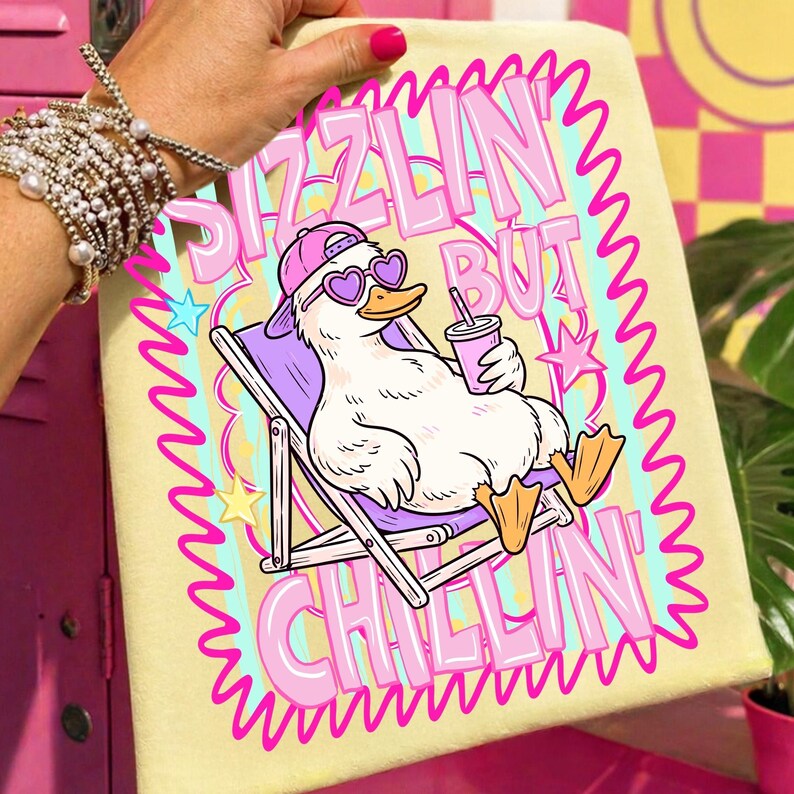 Op de afbeelding: Een lichtgele handdoek met een cartoon eend met een hartvormige zonnebril en een roze pet, ontspannen in een strandstoel. De handdoek heeft de tekst "Sizzlin' But Chillin'" in roze en paars. De eend houdt een roze drankje vast.