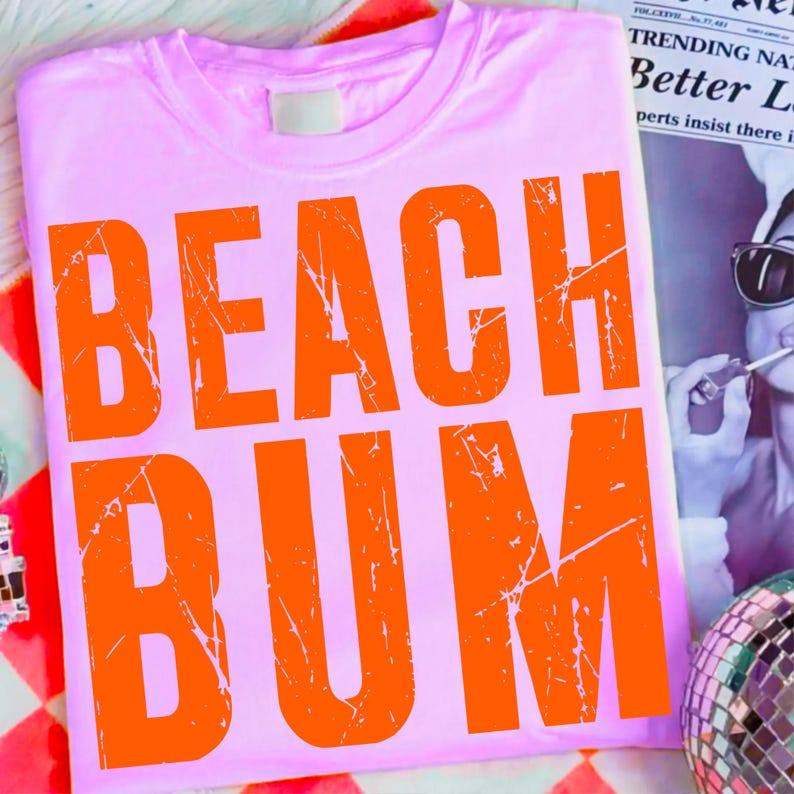 May include: A light pink t-shirt with the words "BEACH BUM" printed in large, distressed orange letters. The shirt is folded and lies on a colorful patterned surface, with a newspaper and disco ball in the background.