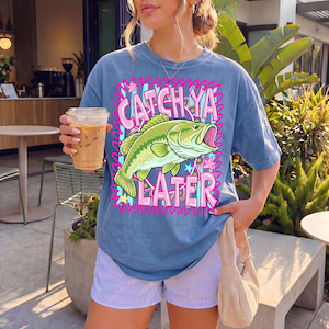 Op de afbeelding: Blauw T-shirt met de tekst "Catch Ya Later" in roze en een groene visafbeelding. Het shirt wordt gedragen met gestreepte shorts en een beige handtas. De persoon houdt een doorzichtige plastic beker met ijskoffie vast.
