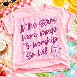 May include: A light pink t-shirt with rolled-up sleeves. The shirt features the text "If The Stars Were Made To Worship So Will I" in a purple, handwritten style, with star accents. The shirt is on a red and white checkered surface.