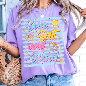 Peut inclure: Un t-shirt lilas avec l'inscription "Sun, Salt, and Sand" dans une police de caract&egrave;res color&eacute;e et ludique. Le motif comprend un coquillage, des n&oelig;uds et des &eacute;toiles de mer. Le t-shirt a une coupe d&eacute;contract&eacute;e et des manches courtes.