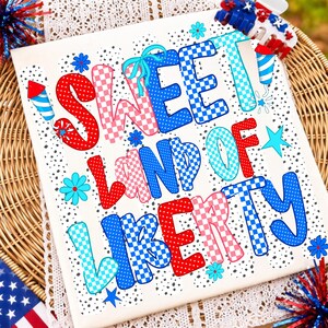 May include: A white t-shirt featuring the text SWEET LAND OF LIBERTY in red, white, and blue checkered and polka dot patterns. The design includes small blue flowers, stars, and firework graphics scattered across the fabric surface.