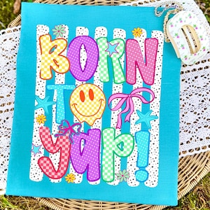 May include: A turquoise fabric featuring the phrase "BORN TO YAP!" in vibrant, patterned lettering. The design incorporates a smiley face, bows, stars, and flowers. The background has white stripes with polka dots.