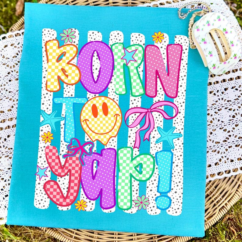 May include: A turquoise fabric featuring the phrase "BORN TO YAP!" in vibrant, patterned lettering. The design incorporates a smiley face, bows, stars, and flowers. The background has white stripes with polka dots.