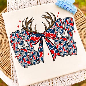 Peut inclure: T-shirt cr&egrave;me avec le mot "GIRL" en motif camouflage rouge, blanc et bleu. Les lettres sont bord&eacute;es de noir et surmont&eacute;es de bois de cerf marron et d'un n&oelig;ud assorti. Des &eacute;toiles rouges ornent le design.