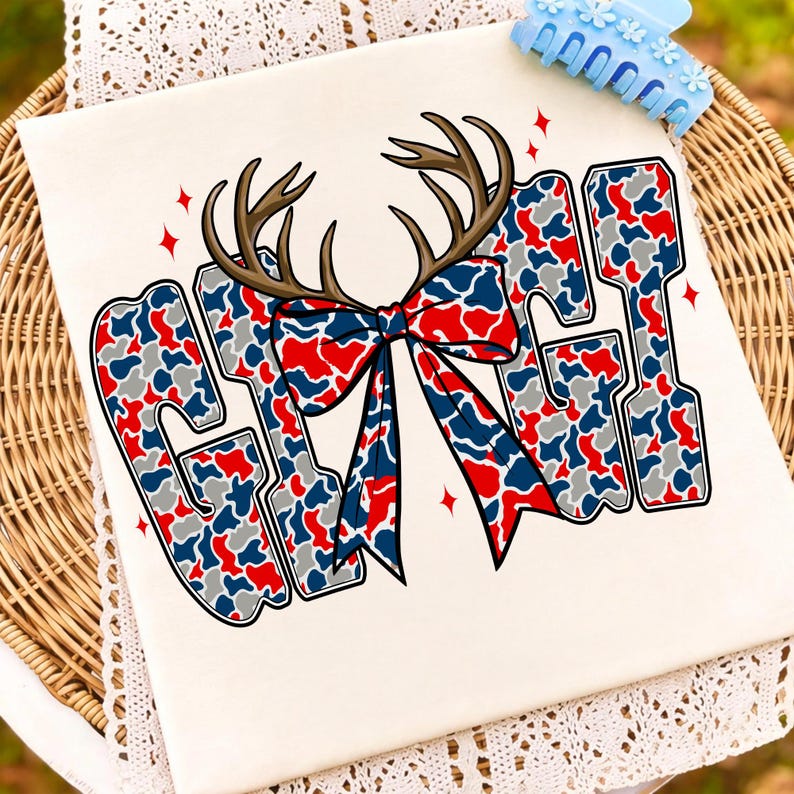 Peut inclure: T-shirt cr&egrave;me avec le mot "GIRL" en motif camouflage rouge, blanc et bleu. Les lettres sont bord&eacute;es de noir et surmont&eacute;es de bois de cerf marron et d'un n&oelig;ud assorti. Des &eacute;toiles rouges ornent le design.