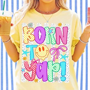 May include: A yellow t-shirt featuring the text "BORN TO YAP!" in a vibrant, multi-coloured design with checkered and polka-dot patterns. The design includes a smiley face, bows, and stars. The shirt is a casual style.
