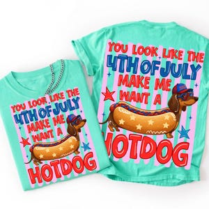 May include: Two turquoise t-shirts with a graphic design. The design features a cartoon dachshund wearing sunglasses and a hat, shaped like a hotdog. Text reads "YOU LOOK LIKE THE 4TH OF JULY MAKE ME WANT A HOTDOG."
