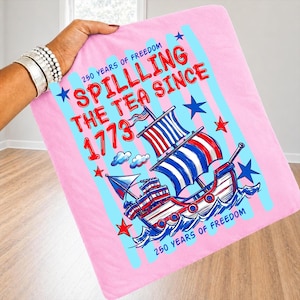 Pu&ograve; includere: Coperta rosa con un'illustrazione nautica di una nave con vele rosse, bianche e blu. La coperta presenta il testo "Spilling the Tea Since 1773" e "250 Years of Freedom" in rosso e blu.