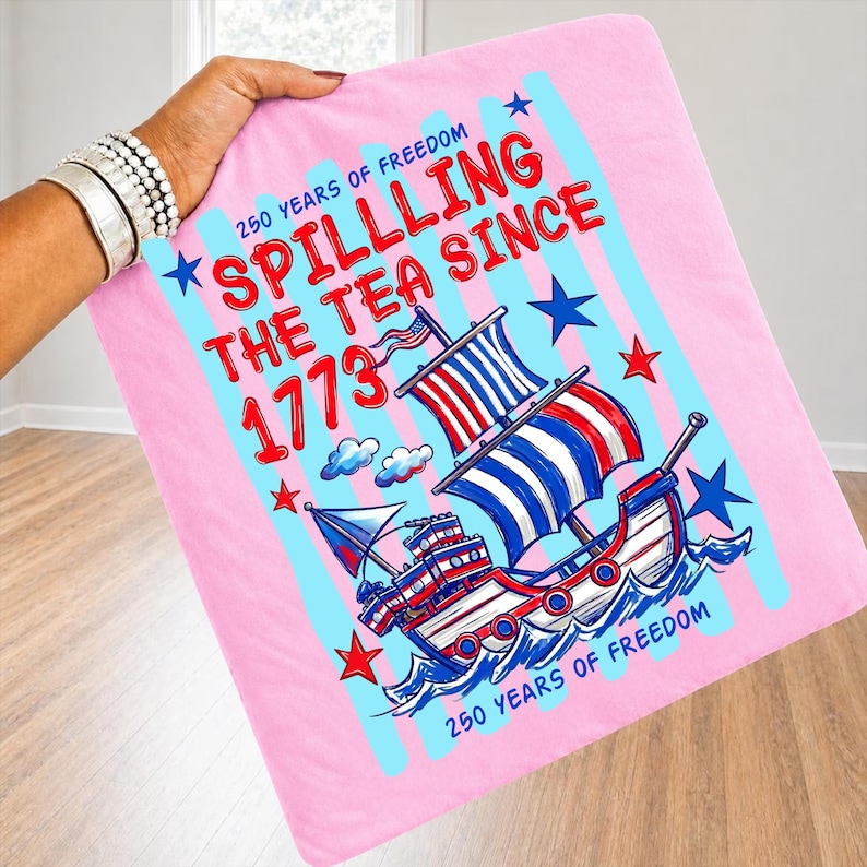 Pu&ograve; includere: Coperta rosa con un'illustrazione nautica di una nave con vele rosse, bianche e blu. La coperta presenta il testo "Spilling the Tea Since 1773" e "250 Years of Freedom" in rosso e blu.