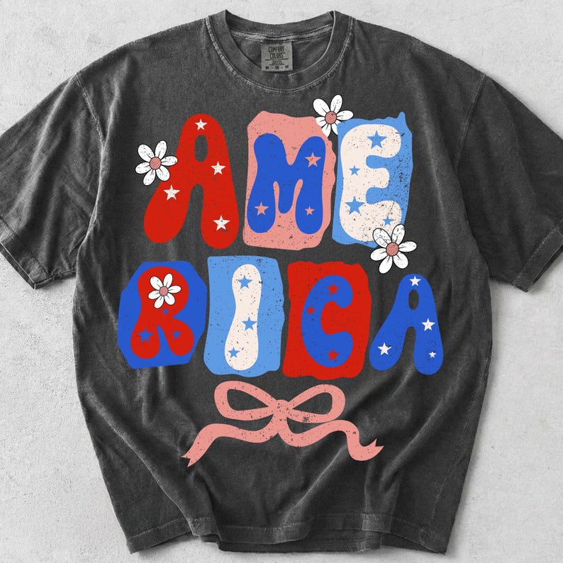 May include: Dark grey t-shirt featuring the word "AMERICA" in red, blue, and pink block letters. The letters are decorated with white stars and flowers. A pink ribbon bow is at the bottom of the design.