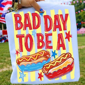K&ouml;nnte beinhalten: Hellblaues T-Shirt mit dem roten Schriftzug "BAD DAY TO BE A". Das Design zeigt zwei Hotdogs mit gelbem Senf, blauen und roten Details und gelben vertikalen Streifen im Hintergrund.