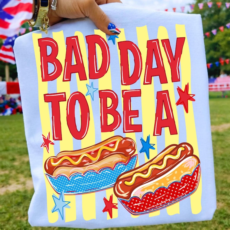 K&ouml;nnte beinhalten: Hellblaues T-Shirt mit dem roten Schriftzug "BAD DAY TO BE A". Das Design zeigt zwei Hotdogs mit gelbem Senf, blauen und roten Details und gelben vertikalen Streifen im Hintergrund.