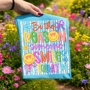 May include: A light blue t-shirt with the text "Be the reason someone smiles today" in colorful, playful lettering. The design includes smiley faces and floral accents. The shirt is held up in front of a colorful flower garden.
