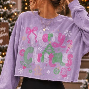 May include: A light purple long-sleeve cropped sweatshirt featuring a graphic print with the text Merry Christmas in pink and green wavy lettering. The design includes illustrations of a bow, a Christmas tree, a candy cane, a gingerbread man, a mitten, and snowflakes.