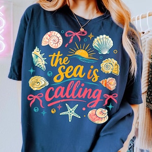 K&ouml;nnte beinhalten: Marineblaues T-Shirt mit einem farbenfrohen Grafikdesign mit Muscheln, einer Sonne und dem Text "the sea is calling". Das Design beinhaltet einen Seestern und Schleifen. Das Shirt ist ein l&auml;ssiger, bequemer Stil.