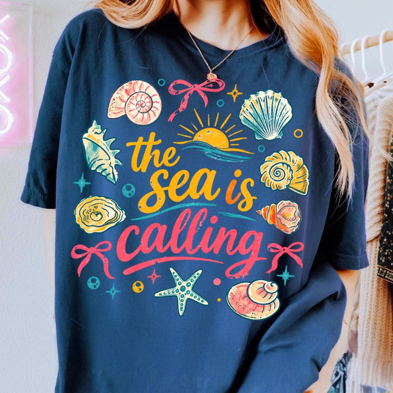 K&ouml;nnte beinhalten: Marineblaues T-Shirt mit einem farbenfrohen Grafikdesign mit Muscheln, einer Sonne und dem Text "the sea is calling". Das Design beinhaltet einen Seestern und Schleifen. Das Shirt ist ein l&auml;ssiger, bequemer Stil.
