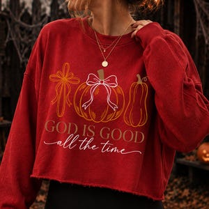 Puede incluir: Una sudadera roja corta de manga larga con un gr&aacute;fico de una cruz, una calabaza con un lazo blanco y una calabaza alargada. El texto "GOD IS GOOD" est&aacute; impreso en letras marrones, con "all the time" en letra cursiva blanca debajo.