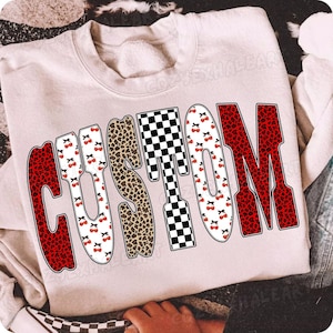 May include: A light pink sweatshirt with the word "CUSTOM" in a bold, playful font. The letters feature a mix of patterns: red leopard print, black and white checkerboard, and cherries. The sweatshirt is casual and comfortable.