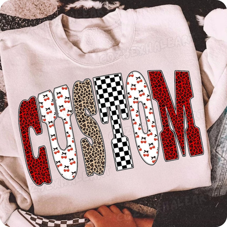 May include: A light pink sweatshirt with the word "CUSTOM" in a bold, playful font. The letters feature a mix of patterns: red leopard print, black and white checkerboard, and cherries. The sweatshirt is casual and comfortable.