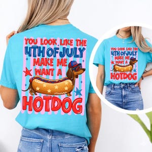 May include: Light blue t-shirt with a graphic featuring a dachshund wearing sunglasses and a hat, designed as a hotdog. The text reads "YOU LOOK LIKE THE 4TH OF JULY MAKE ME WANT A HOTDOG".