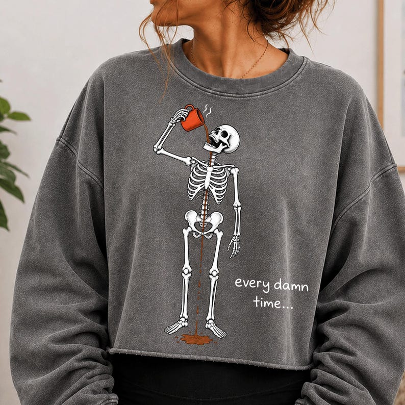 May include: A charcoal grey crewneck sweatshirt featuring a graphic of a white skeleton drinking from an orange mug. The coffee flows through the skeleton's ribcage and spills onto the floor. The text "every damn time..." is printed in white.