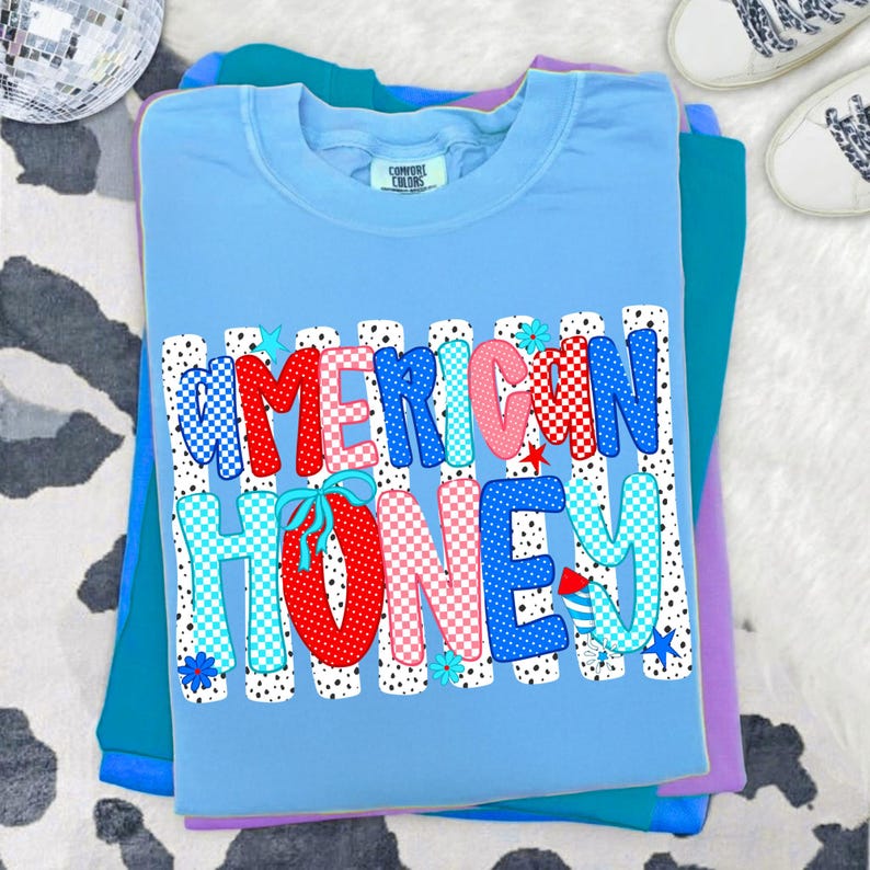 May include: A light blue crew neck t-shirt featuring the text AMERICAN HONEY in a bold, multi-colored, patterned font. The letters are decorated with red, blue, and pink polka dots and checkered patterns, accented by small star and flower icons.