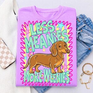 May include: A lilac-colored t-shirt featuring a brown dachshund dog illustration. The shirt has the text "LESS MEANIES MORE WEENIES" in a colorful, playful font. The design includes stars and wavy lines, creating a fun, eye-catching graphic.