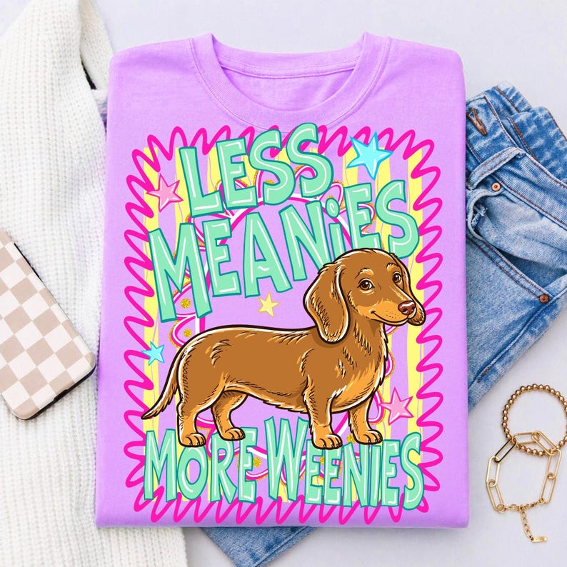 May include: A lilac-colored t-shirt featuring a brown dachshund dog illustration. The shirt has the text "LESS MEANIES MORE WEENIES" in a colorful, playful font. The design includes stars and wavy lines, creating a fun, eye-catching graphic.