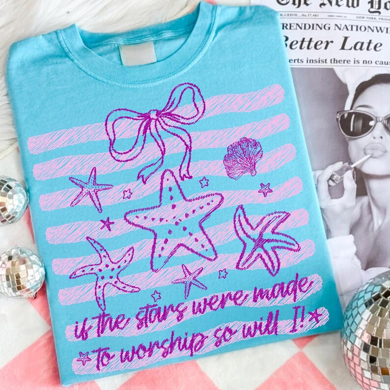 Op de afbeelding: Lichtblauw T-shirt met een ontwerp van zeesterren, een strik en een zeeschelp in roze en paars. Het shirt heeft roze horizontale strepen en de tekst "If the stars were made to worship so will I!"