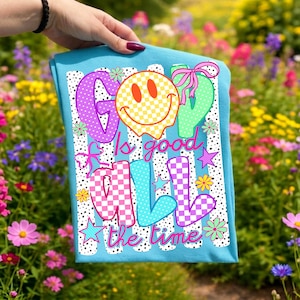 Op de afbeelding: Een felblauw t-shirt met een grafische print met de tekst "GOD is good ALL the time". Het ontwerp bevat een geel geblokt smileygezicht, kleurrijke letters met ruitpatronen, kleine bloemen, sterren en roze strikjes.