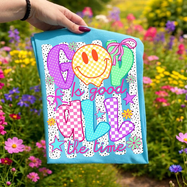 Op de afbeelding: Een felblauw t-shirt met een grafische print met de tekst "GOD is good ALL the time". Het ontwerp bevat een geel geblokt smileygezicht, kleurrijke letters met ruitpatronen, kleine bloemen, sterren en roze strikjes.