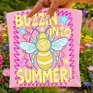 K&ouml;nnte beinhalten: Rosafarbenes Grafik-T-Shirt mit einer Cartoon-Bienen-Illustration. Das T-Shirt tr&auml;gt den Text "BUZZIN' INTO SUMMER!" in gelben Blockbuchstaben. Die Biene ist gelb und blau, mit einem rosa-gelben Hintergrund.