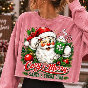 K&ouml;nnte beinhalten: Ein altrosa Langarm-Sweatshirt mit einer Grafik vom zwinkernden Weihnachtsmann, der einen gr&uuml;nen Becher Kakao mit Zuckerstange h&auml;lt. Der Text lautet Cozy & Merry SANTA'S COCOA CLUB, umgeben von Stechpalmen, Tannenb&auml;umen und roten Weihnachtskugeln.