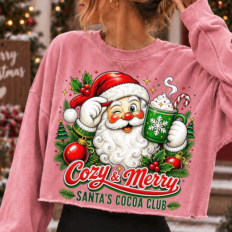 K&ouml;nnte beinhalten: Ein altrosa Langarm-Sweatshirt mit einer Grafik vom zwinkernden Weihnachtsmann, der einen gr&uuml;nen Becher Kakao mit Zuckerstange h&auml;lt. Der Text lautet Cozy & Merry SANTA'S COCOA CLUB, umgeben von Stechpalmen, Tannenb&auml;umen und roten Weihnachtskugeln.