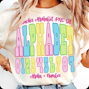 May include: Cream-coloured t-shirt with a vibrant alphabet and number design. The design features the word "ALPHABET" in large, vertically striped letters, and the numbers 0 to 9. The text "sucker Alphabet PNG set" and "Alpha + Number" are also included.