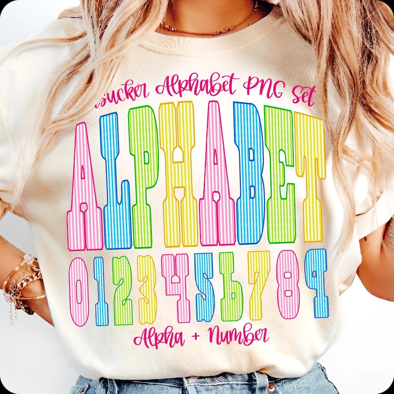 May include: Cream-coloured t-shirt with a vibrant alphabet and number design. The design features the word "ALPHABET" in large, vertically striped letters, and the numbers 0 to 9. The text "sucker Alphabet PNG set" and "Alpha + Number" are also included.