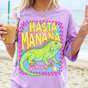 K&ouml;nnte beinhalten: Ein lila T-Shirt mit einer gro&szlig;en Grafik einer gr&uuml;nen Leguan und dem gelben Text "HASTA MANANA". Der Leguan hat blaue und gr&uuml;ne Streifen. Der Text ist rosa und blau umrandet, mit Sternen und Wellenlinien.
