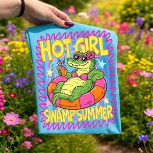 May include: A light blue t-shirt featuring a cartoon alligator in a pink bikini and sunglasses, relaxing on an orange and pink inflatable ring. The text "HOT GIRL SWAMP SUMMER" is displayed in yellow and pink.