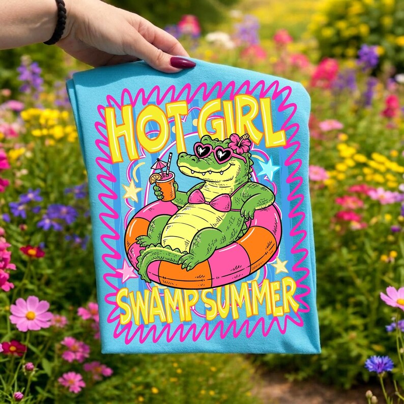 May include: A light blue t-shirt featuring a cartoon alligator in a pink bikini and sunglasses, relaxing on an orange and pink inflatable ring. The text "HOT GIRL SWAMP SUMMER" is displayed in yellow and pink.