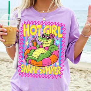 May include: A lilac-colored t-shirt with a graphic of an alligator in a pink bikini and sunglasses, floating in a pink and orange inner tube. The text reads "HOT GIRL SWAMP SUMMER" in yellow and pink.