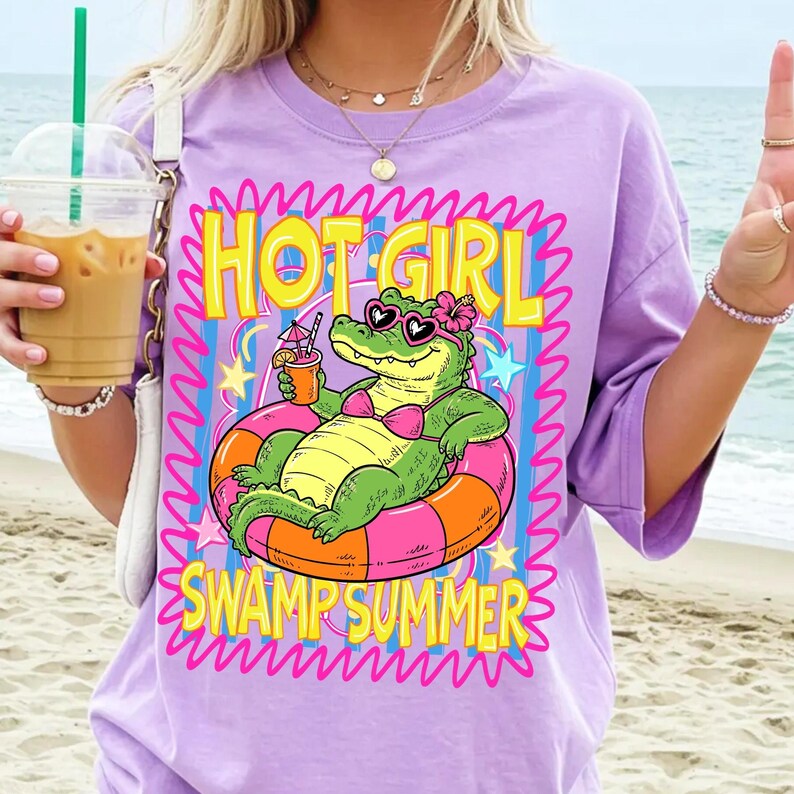 May include: A lilac-colored t-shirt with a graphic of an alligator in a pink bikini and sunglasses, floating in a pink and orange inner tube. The text reads "HOT GIRL SWAMP SUMMER" in yellow and pink.
