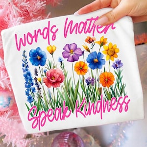 Puede incluir: Bolso tote blanco con un dise&ntilde;o floral y el texto rosa "Words Matter Speak Kindness". El arreglo floral incluye flores azules, amarillas, naranjas y moradas con tallos y hojas verdes.