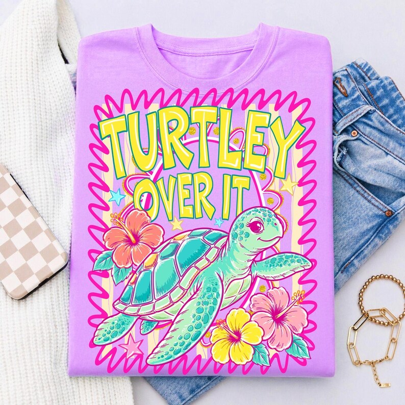 Puede incluir: Una camiseta de color lila con un dise&ntilde;o de tortuga de dibujos animados y el texto "TURTLEY OVER IT". El dise&ntilde;o incluye una tortuga turquesa, flores de hibisco rosas y amarillas, y un borde ondulado rosa. La camiseta se muestra con vaqueros azules y una pulsera dorada.