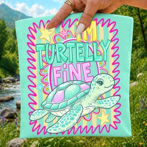 May include: A light teal tote bag with a cartoon turtle illustration and the text "I'm Turtelly Fine". The turtle has a teal shell with pink accents and is surrounded by stars and a pink zig-zag border.