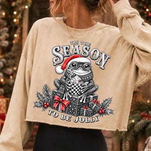 Puede incluir: Una sudadera corta de color beige con el gr&aacute;fico de una rana con gorro de Pap&aacute; Noel y gafas, sosteniendo una taza de cacao sobre regalos. El texto dice 'TIS THE SEASON TO BE JOLLY' con detalles de hojas de acebo y bayas rojas.