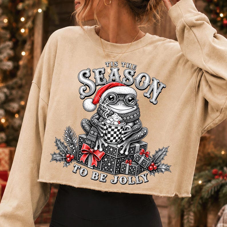 Puede incluir: Una sudadera corta de color beige con el gr&aacute;fico de una rana con gorro de Pap&aacute; Noel y gafas, sosteniendo una taza de cacao sobre regalos. El texto dice 'TIS THE SEASON TO BE JOLLY' con detalles de hojas de acebo y bayas rojas.