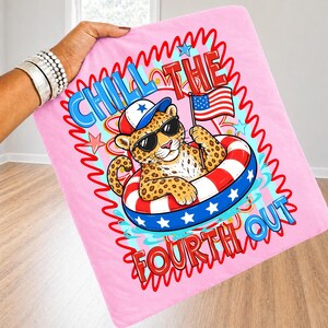 Op de afbeelding: Roze deken met een cartoonluipaard met een zonnebril en een patriotische hoed, drijvend in een rood, wit en blauw zwemband. De deken heeft de tekst "Chill the Fourth Out" met een Amerikaanse vlag.